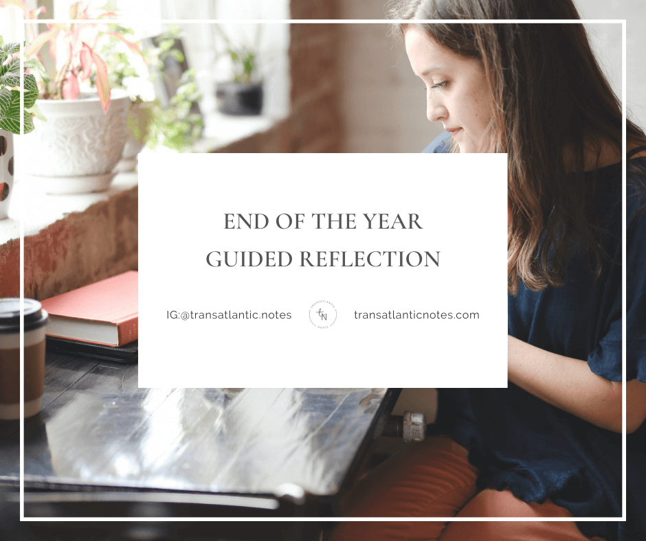 Foreground Text: End of the Year Guided Reflection Tag; Background Image: A woman sits at a long, black wooden desk near a large window. She’s writing in a notebook, reflecting on the past year. Photo by Hannah Olinger via Unsplash.