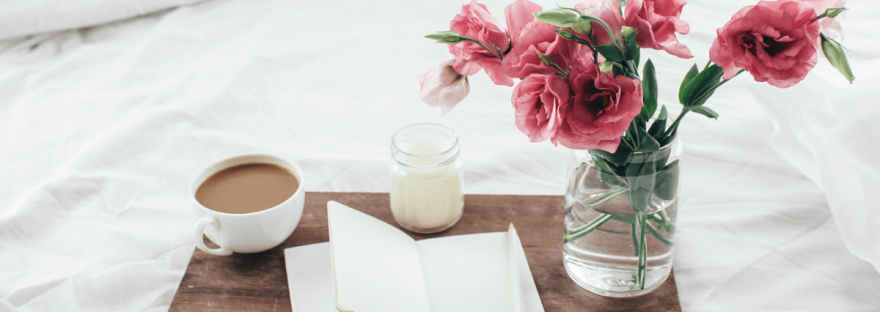 Wooden tray with paper sketchbook, candle and spring flowers on clean white bedding. Photo by Alena Ozerova via Canva.