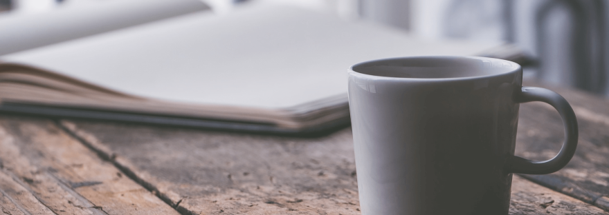 On a worn wooden table is an open journal (notebook) and a white ceramic coffee mug; by Jessica Lewis via Unsplash.