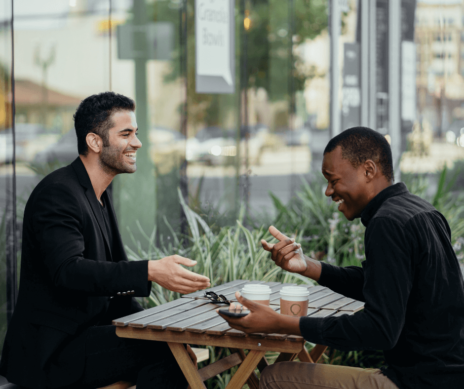Two male friends sit opposite each other at a table drinking coffee; one is smiling/laughing as he listens to what the other is saying. Photo by RODNAE Productions via Pexels.