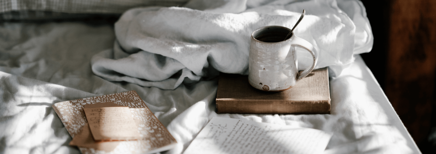 A collection of notebooks and a grey mug on a bed; photo via Annie Spratt/Unsplash