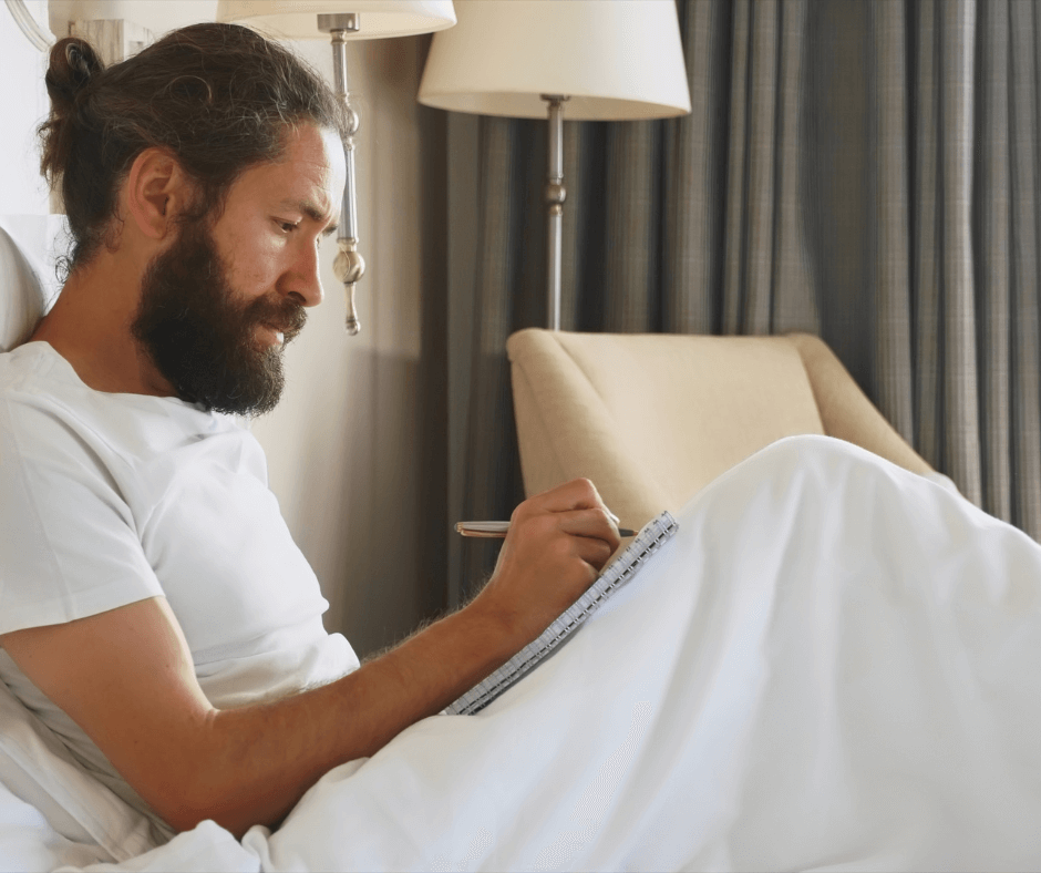 A young bearded man sits in his bed writing notes in his diary/journal.