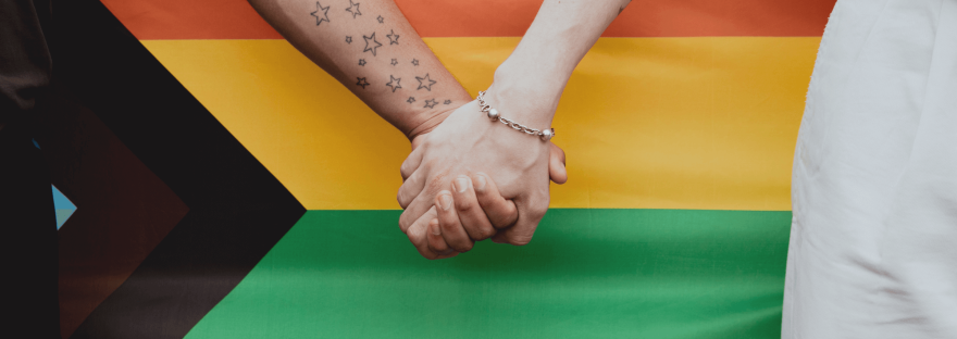 Two people holding hands in front of a progress Pride flag; photo via Emma Rahmani/Canva.