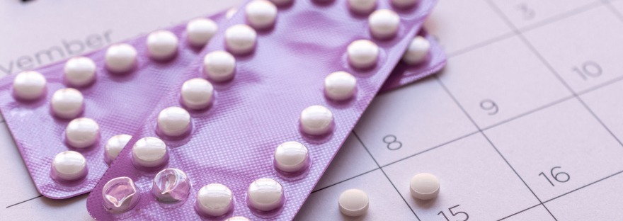 Two packets of birth control pills on top of a monthly calendar; photo via Noel Smiley/Canva