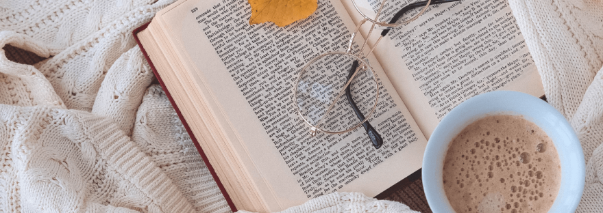 An autumn aesthetic composition shows a cream knitted sweater, an open book, cup of hot chocolate, yellow leaves and small orange pumpkins; photo via Vita Hnatiuk/Canva.