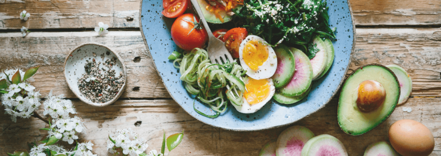 A large blue salad bowl filled to the brim with low-carb foods like kale, sliced eggs, mashed avocado, sliced cucumber and grated carrot; photo via Brooke Lark/[Canva.