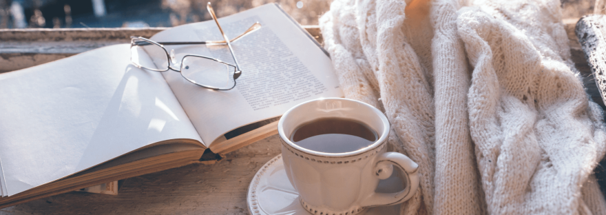 An open book, cup of tea, and white sweater on a window sill bathed in sunlight; photo via Alena Ozerova/Canva.