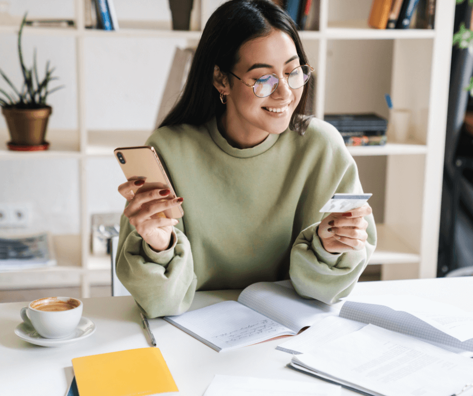 A happy, smiling young woman sits at a desk using a budgeting app on her phone to check the credit card she is holding.