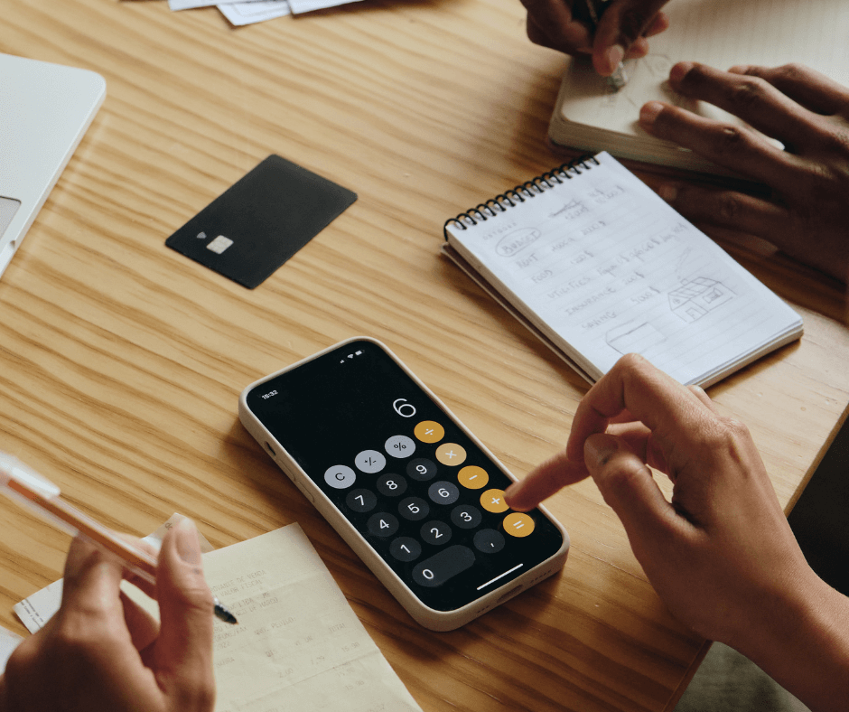 A couple sitting at a table working on their household finances (only their hands are visible); one writes out expense on a notepad while the other uses the calculator app on their mobile phone.