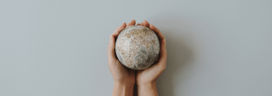 Against a plain, light grey background are the hands of a person carefully holding a small geographical globe; photo via floraldecor/Canva.
