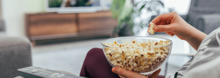 A person sits watching television holding a bowl of popcorn; photo via hsyncoban/Canva.
